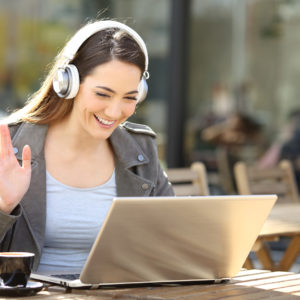Happy woman wearing wireless headset videocalling on laptop sitting in a cafe terrace